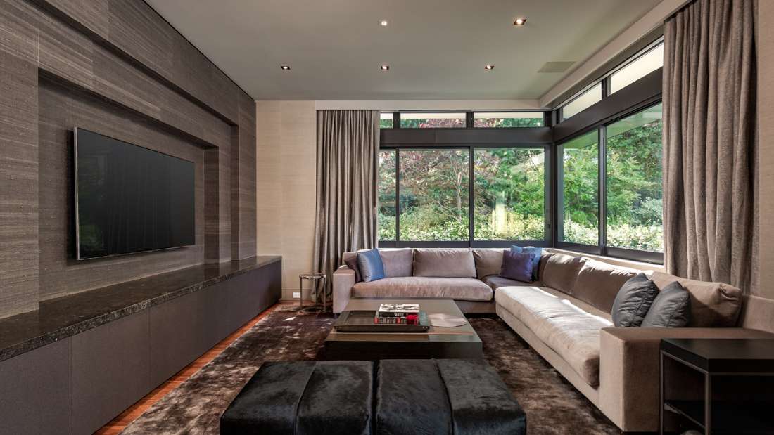 Contemporary living room with large sectional sofa, floor-to-ceiling windows overlooking lush greenery, and sleek entertainment wall with mounted flat-screen TV.