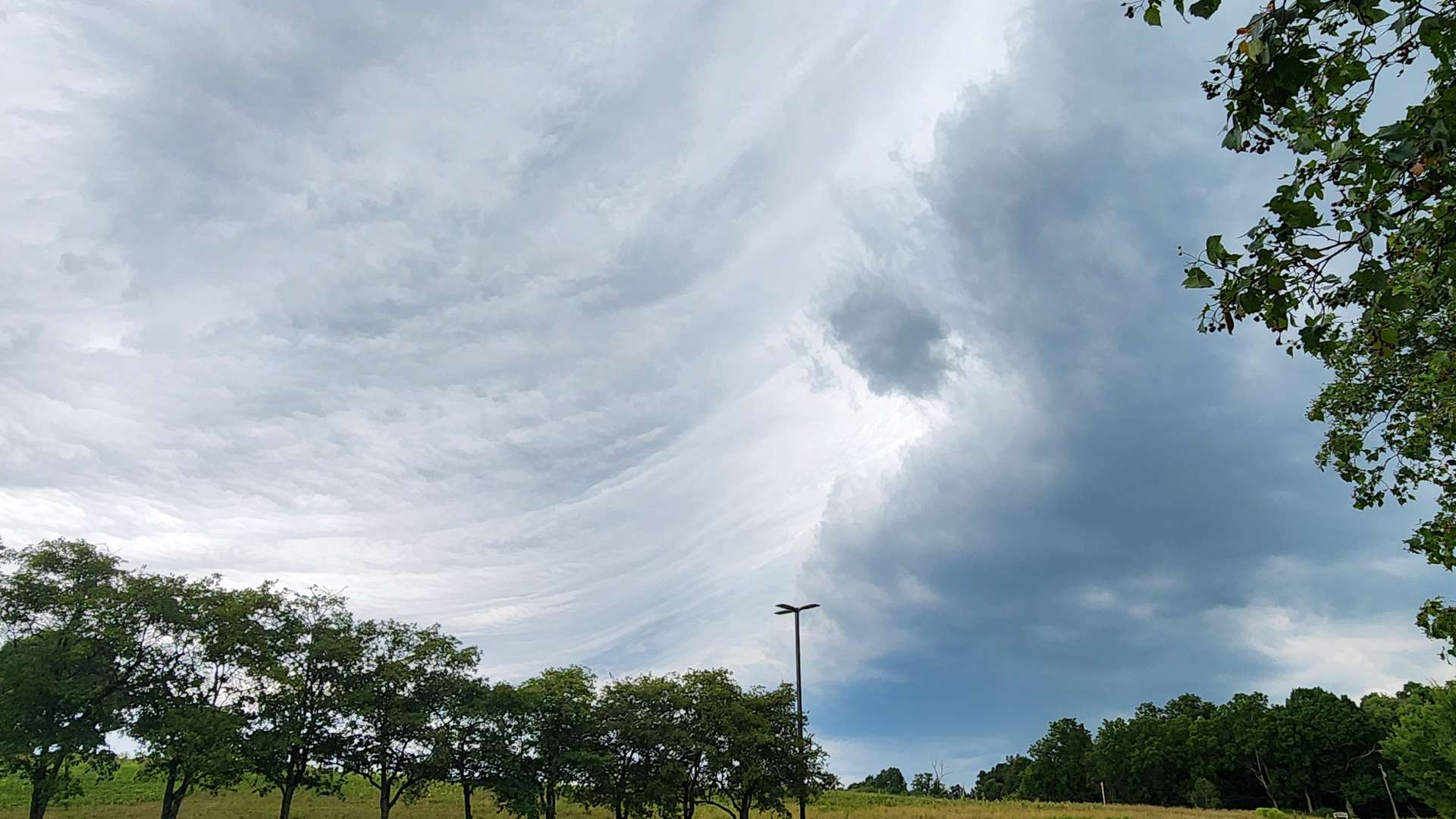 Parking lot with two cars under dramatic swirling storm clouds, bordered by a row of trees and grassy field.