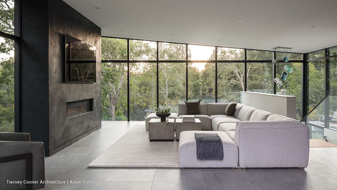 Modern living room with floor-to-ceiling glass walls overlooking lush greenery, gray sectional sofa with matching cushions, sleek black stone fireplace with mounted TV, and minimalist decor featuring neutral tones.