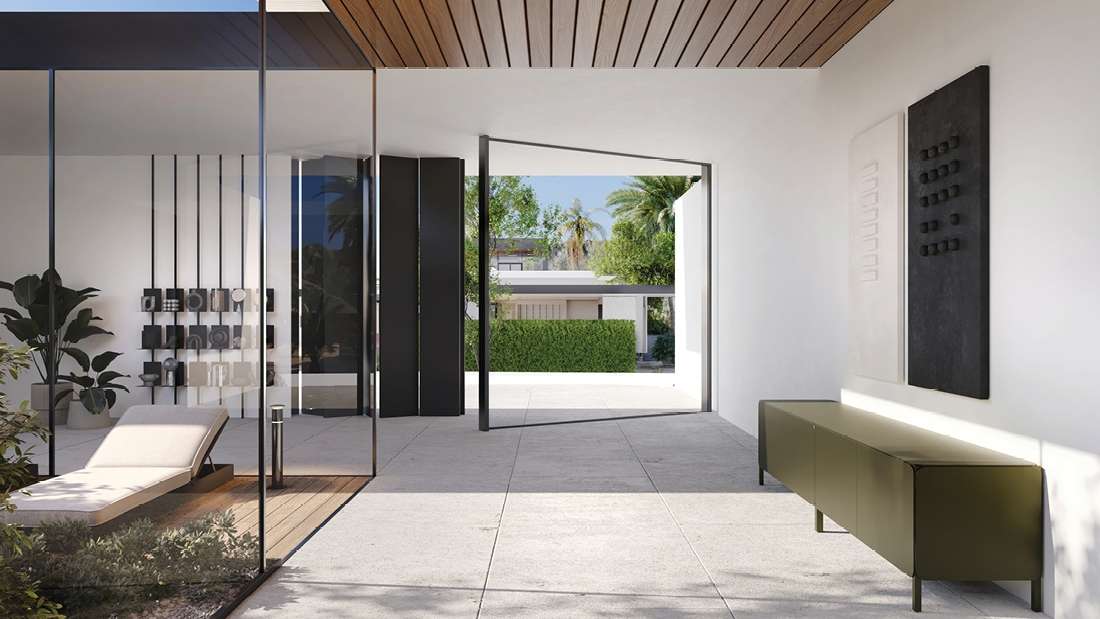 Contemporary home interior with glass walls, minimalist lounge chair, modern art panels, and open doorway leading to a landscaped outdoor area with palm trees.