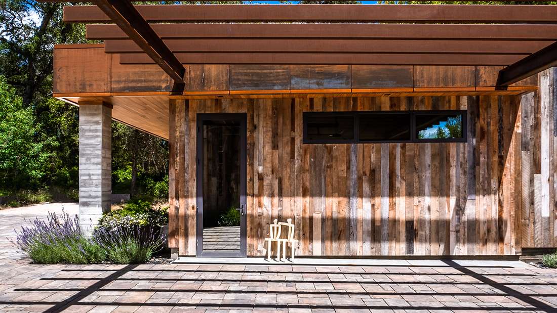 Modern home exterior with reclaimed wood siding, black-framed glass door, horizontal window, and steel pergola casting geometric shadows on stone paver patio.