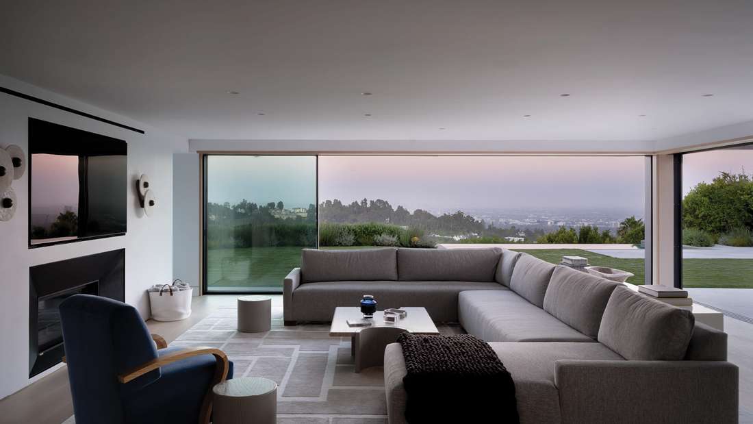 Spacious modern living room with large sectional sofa, floor-to-ceiling glass walls, panoramic garden and city views at sunset.