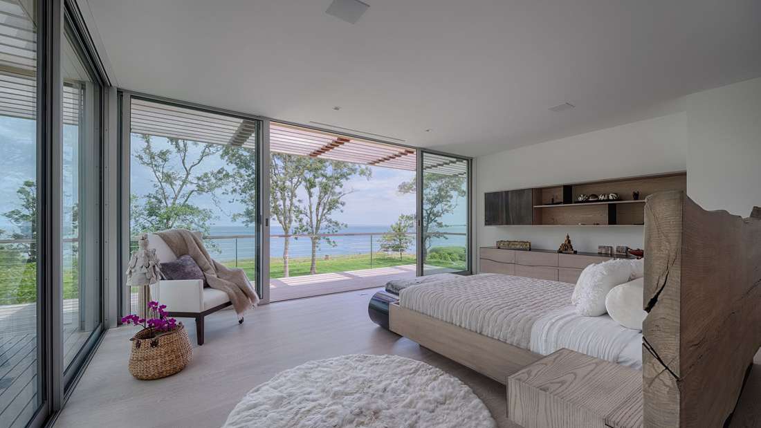 Modern coastal bedroom with floor-to-ceiling glass walls, light wood flooring, minimalist furniture, and panoramic ocean views from a private terrace.