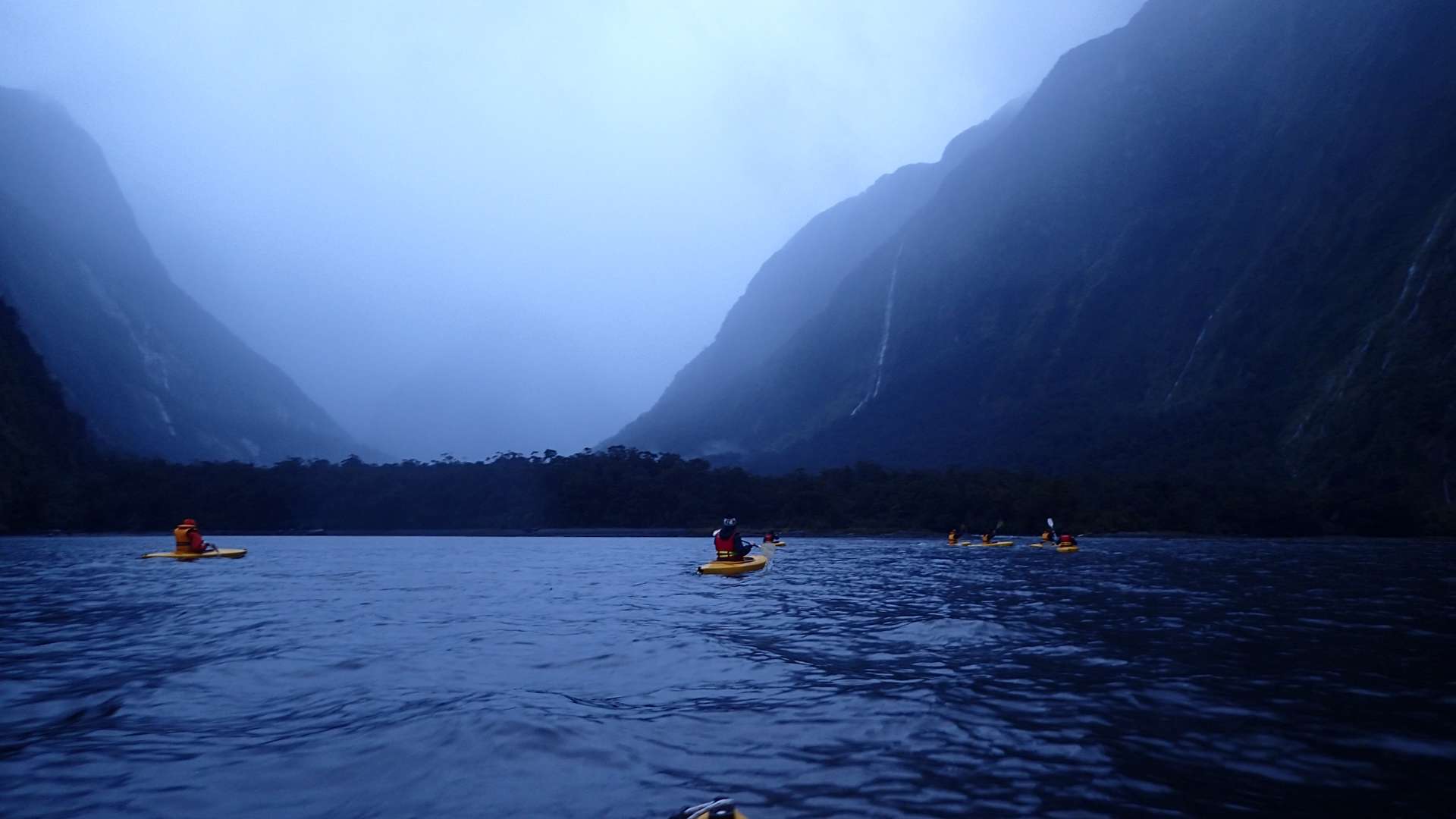 Group of kayakers paddling on a misty blue fjord surrounded by steep dark mountains under overcast skies.