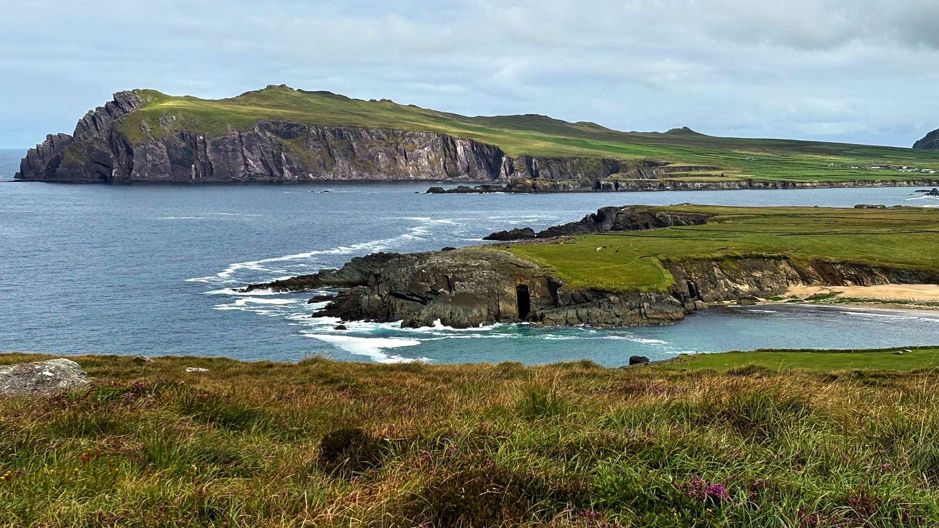Rocky coastline with grassy cliffs and ocean waves under a cloudy sky, featuring a large headland in the distance.