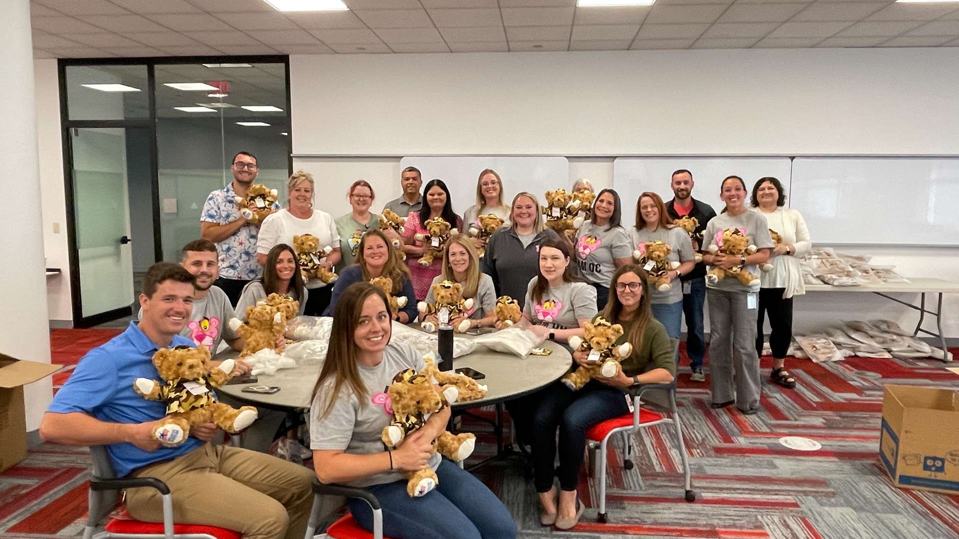 Group of people gathered in a modern office space, sitting and standing around tables holding teddy bears, with boxes and supplies visible in the background on red and gray patterned carpet.