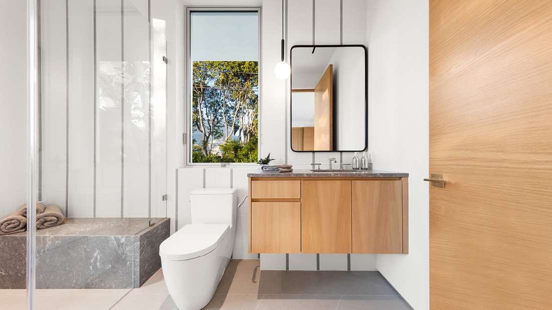 Modern bathroom with frameless glass shower enclosure, gray stone bench, white toilet, floating wood vanity with dark stone countertop, black-framed mirror, and pendant light, featuring a large window with outdoor greenery view.