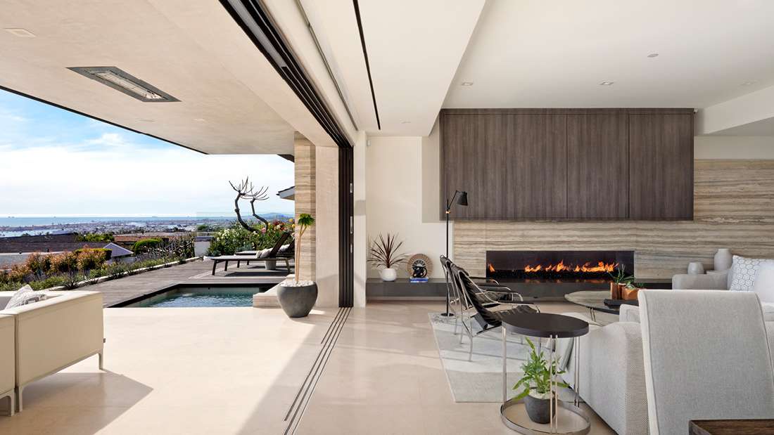 Luxury open-concept living area with modern fireplace, neutral-toned furniture, and sliding glass doors opening to a terrace with pool and panoramic ocean views.