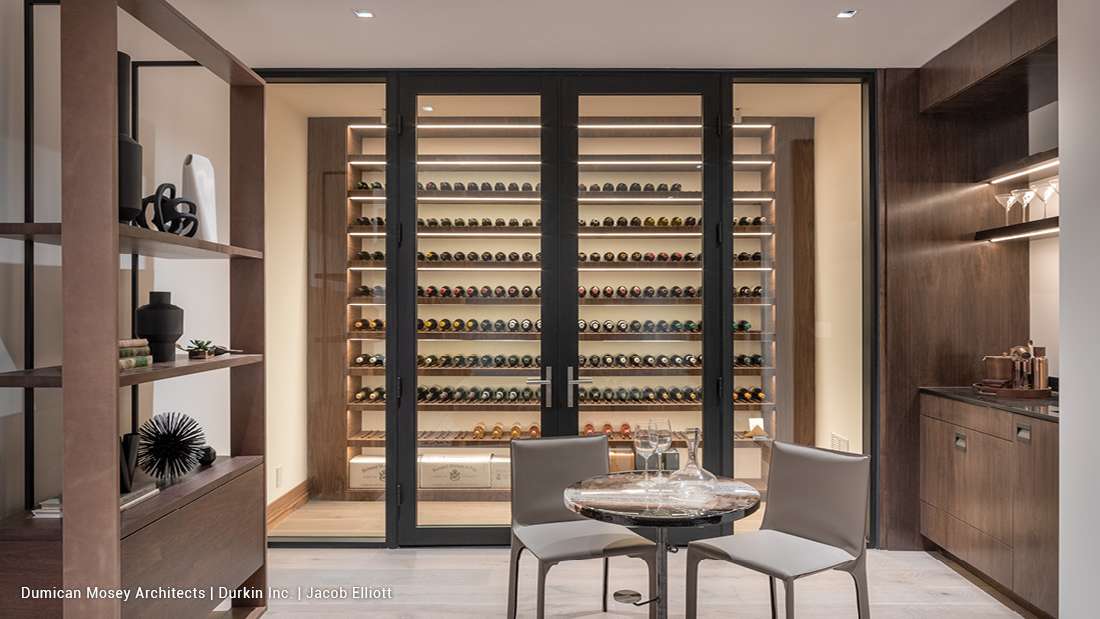 Luxury wine cellar with floor-to-ceiling glass doors, backlit wooden shelves displaying organized wine bottles, and adjacent tasting area featuring round marble table and modern chairs.