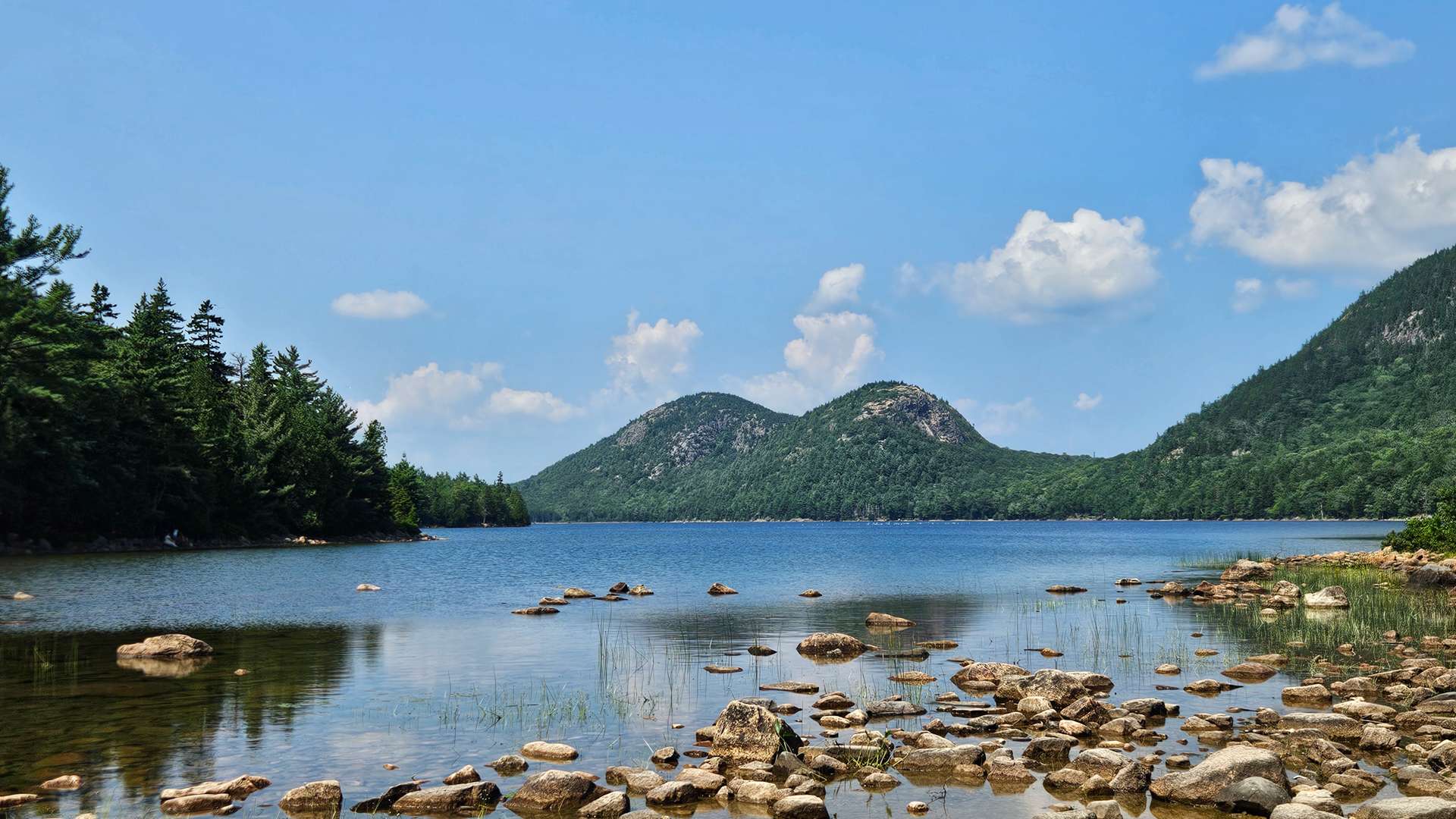 Rocky shoreline of a calm lake with twin rounded mountains in the background under a clear blue sky.