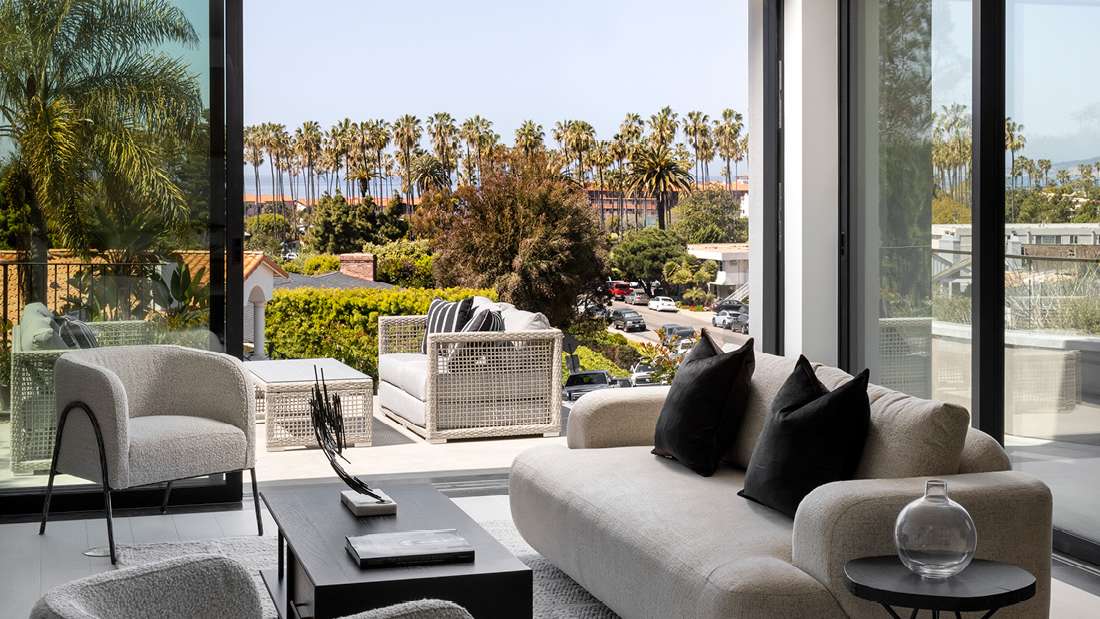 Luxury living room with floor-to-ceiling glass doors opening to a balcony with outdoor seating, beige curved sofa with black accent pillows, modern armchair, and coffee table overlooking palm trees and ocean views.