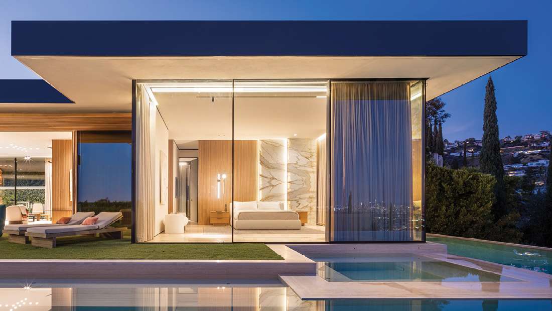 Luxury modern bedroom with floor-to-ceiling glass walls, marble accent wall, private pool, and outdoor lounge area overlooking hillside views at dusk.