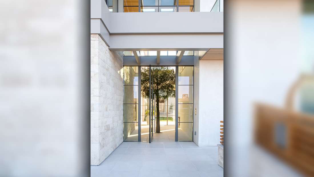 Modern home entryway with double glass pivot doors framed by black metal, floor-to-ceiling windows, and light stone walls under a minimalist overhang with exposed beams.
