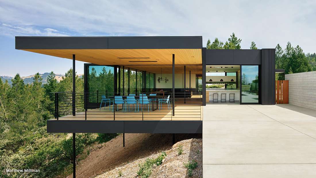 Modern cantilevered home with flat roof, black metal framing, wood-paneled ceiling, and glass walls opening to an outdoor dining area with blue chairs overlooking forested hillside.
