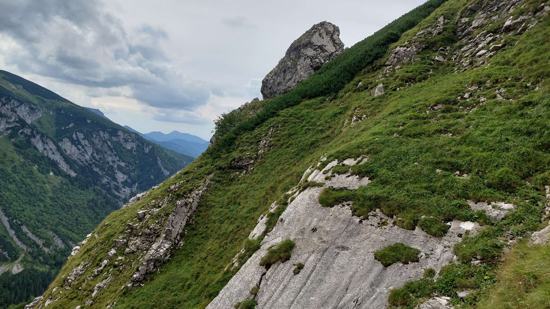 Steep grassy mountain slope with rocky outcrops and distant peaks under a cloudy sky.
