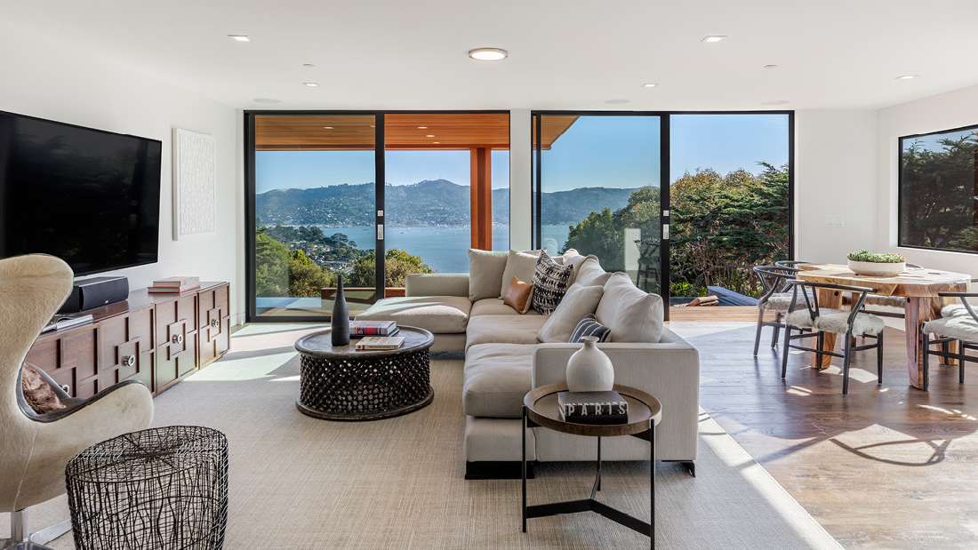 Contemporary living and dining area with large sectional sofa, modern decor, floor-to-ceiling glass doors opening to a balcony with stunning bay and mountain views.