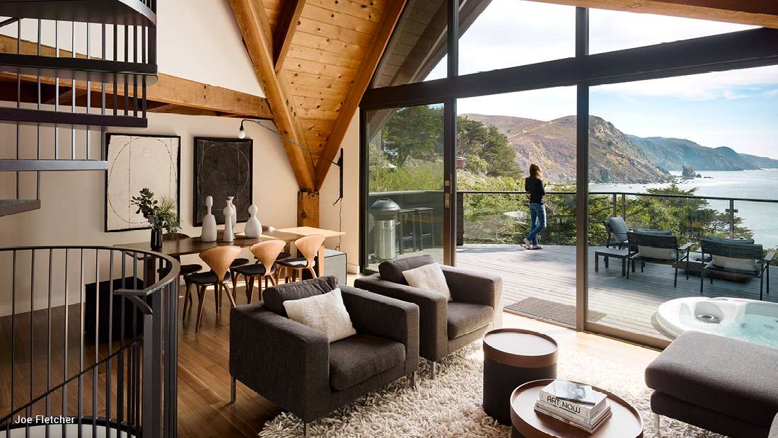 Rustic modern living and dining space with exposed wooden beams, spiral staircase, cozy seating area, and floor-to-ceiling glass doors opening to a deck with ocean and cliff views.