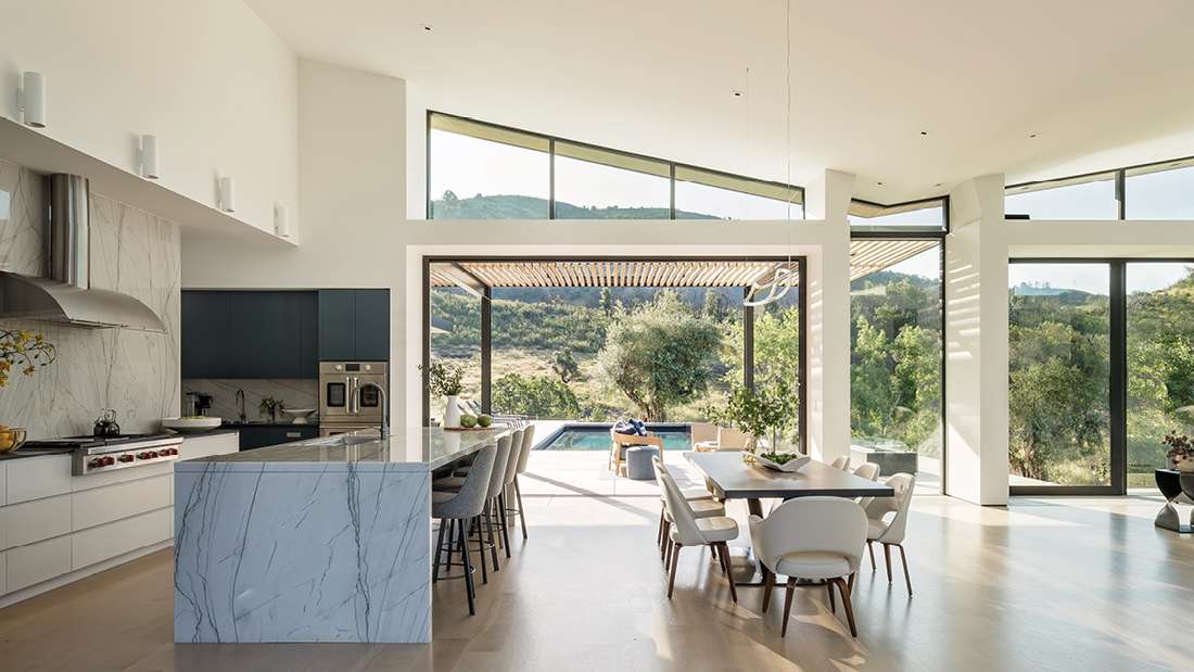 Luxury open-concept kitchen and dining area with marble waterfall island, upholstered barstools, modern dining table with white chairs, and floor-to-ceiling glass walls opening to a patio with slatted pergola and pool overlooking hillside views.