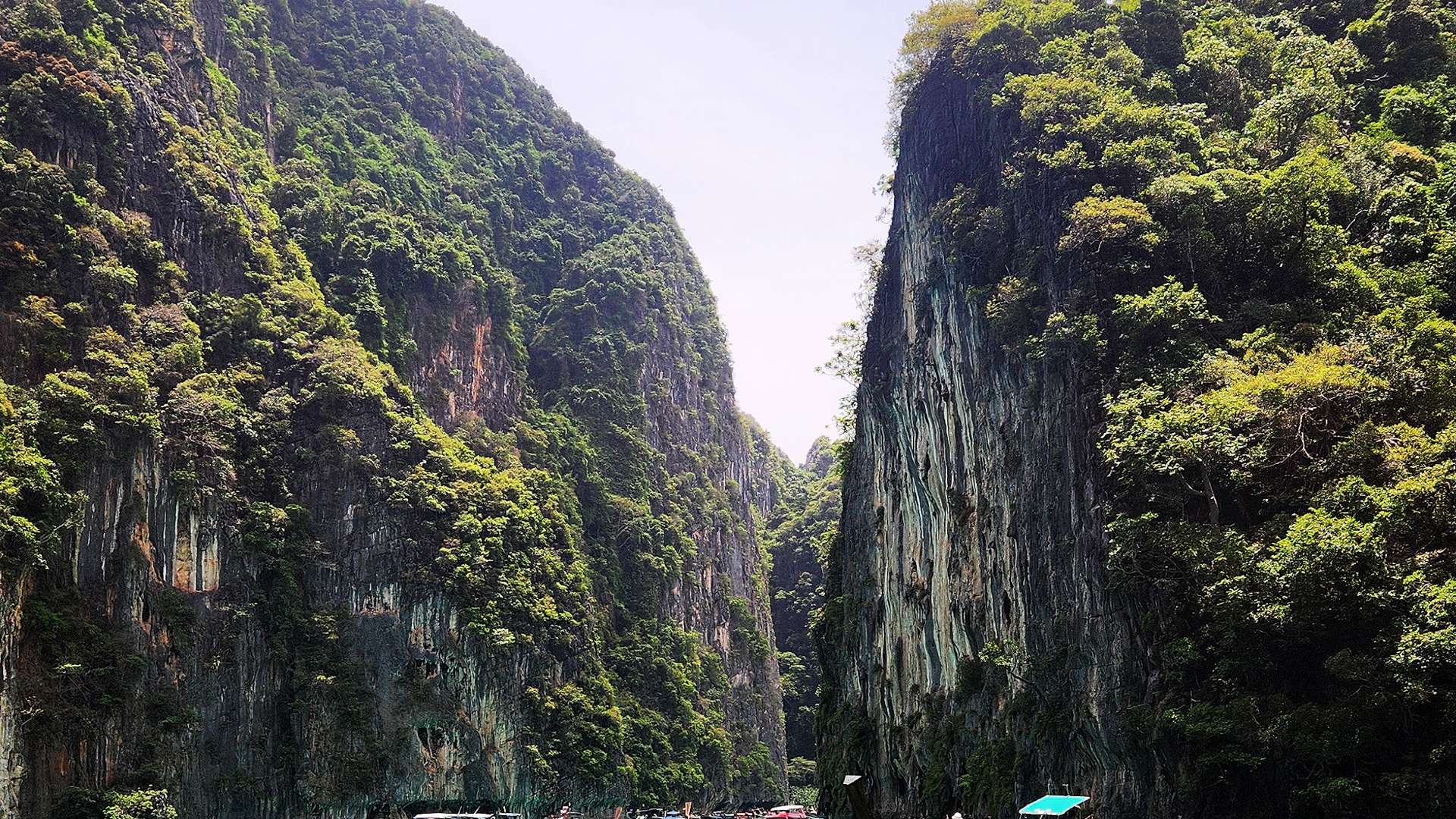 Long-tail boats floating on turquoise water between towering limestone cliffs covered in greenery.