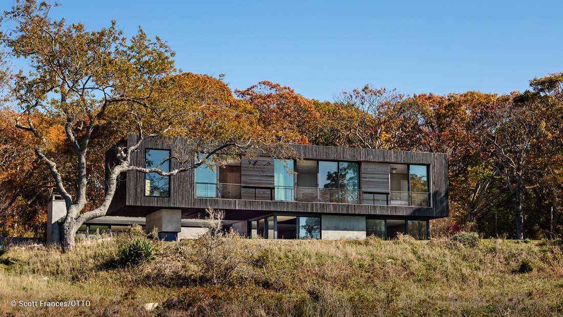 Contemporary home featuring cantilevered design, dark wood siding, expansive floor-to-ceiling glass windows, and elevated structure surrounded by autumn foliage.