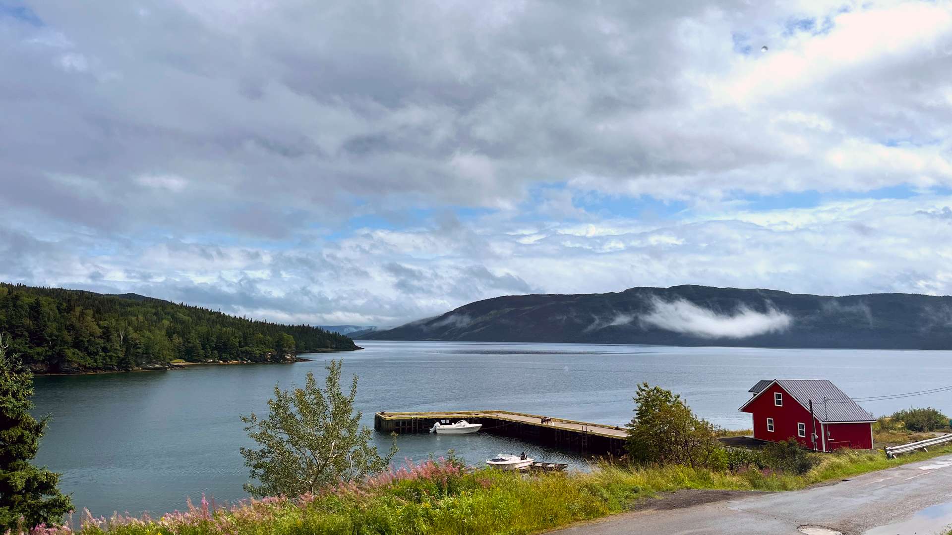 Scenic lakeside view with a wooden dock, small boats, and a bright red house against forested hills and cloudy sky.
