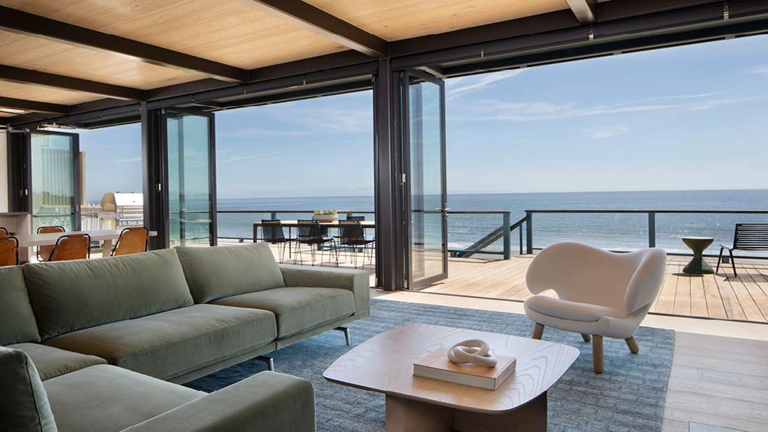 Luxury oceanfront living room with green velvet sectional sofa, sculptural white lounge chair, light wood coffee table, and folding glass doors opening to a spacious deck with panoramic ocean views.