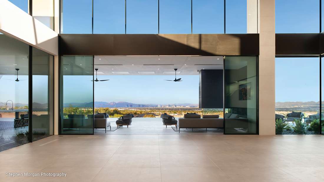 Spacious modern home interior with floor-to-ceiling glass walls, open terrace seating, and panoramic city and mountain views under clear blue skies.