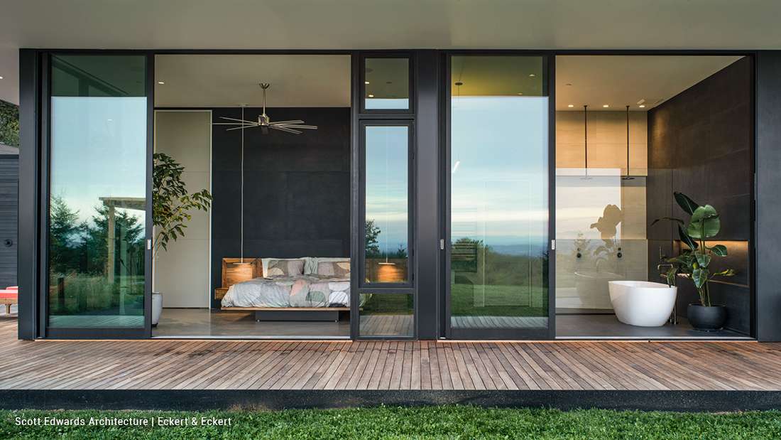 Modern bedroom and bathroom suite with floor-to-ceiling glass walls, minimalist black accent walls, floating bed with pendant lights, and freestanding white bathtub on wooden deck overlooking scenic views.