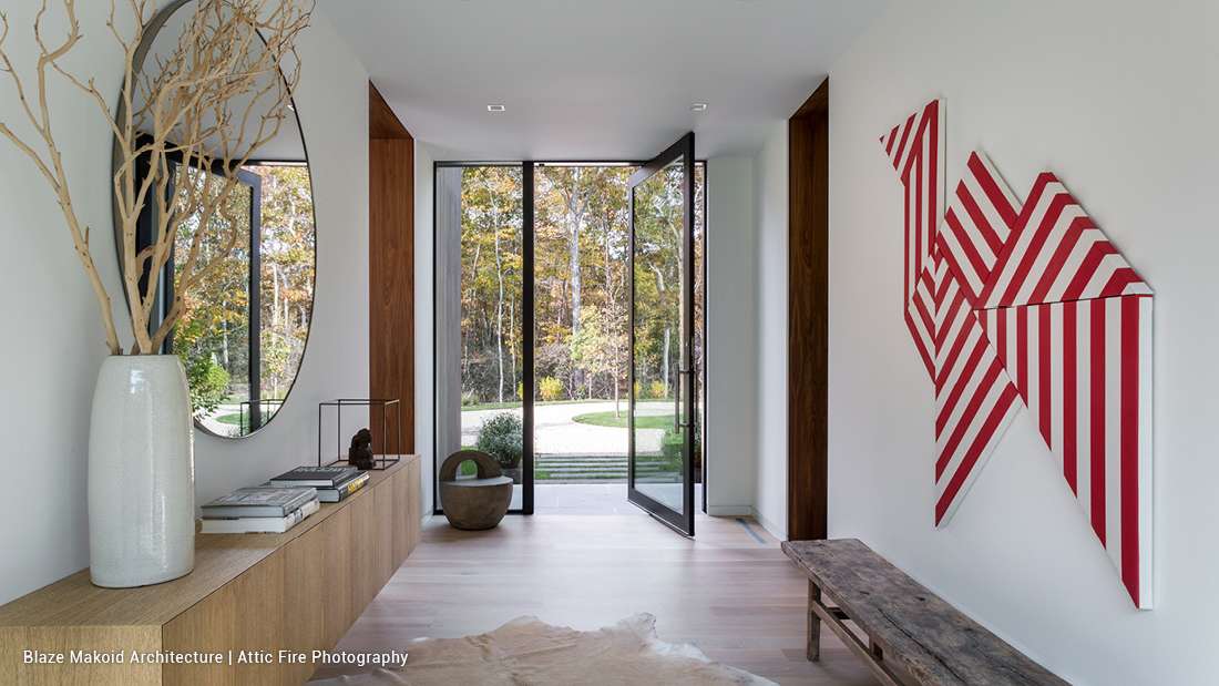 Modern entryway with floor-to-ceiling glass pivot door, minimalist white walls, light wood flooring, natural wood accents, and contemporary decor including round wall mirror, sculptural vase, and geometric red-striped wall art.