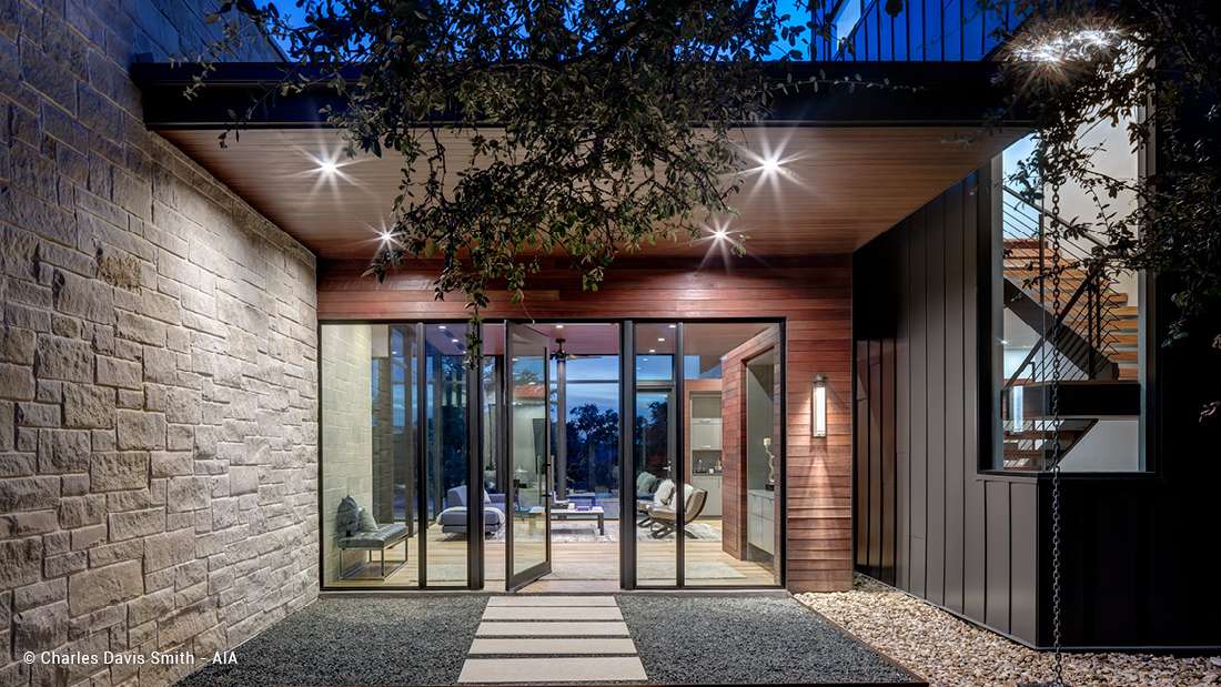 Modern home exterior with mixed materials including stone cladding and vertical wood siding, glass folding doors opening to a living area, and covered entryway with recessed lighting and minimalist landscaping.