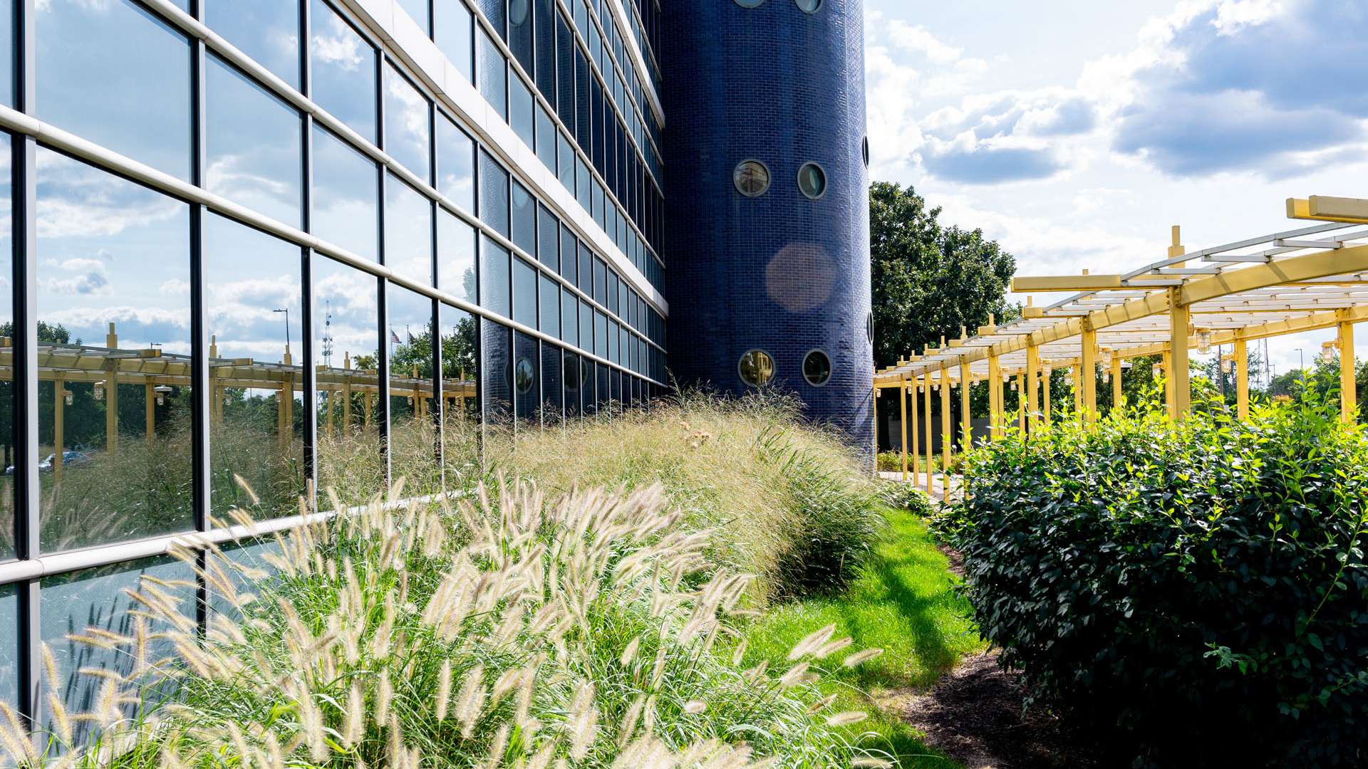 "Reflective glass building with cylindrical blue tower and landscaped greenery under a partly cloudy sky.