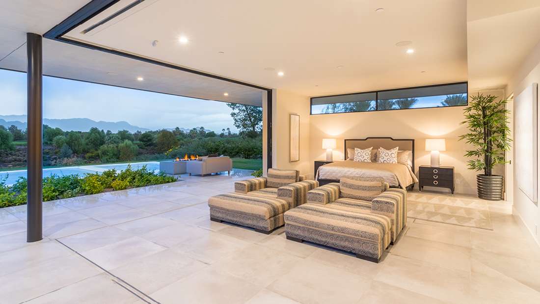 Luxury open-air bedroom with modern furniture, large tile flooring, and panoramic views of mountains and greenery, featuring outdoor lounge area with fire pit.
