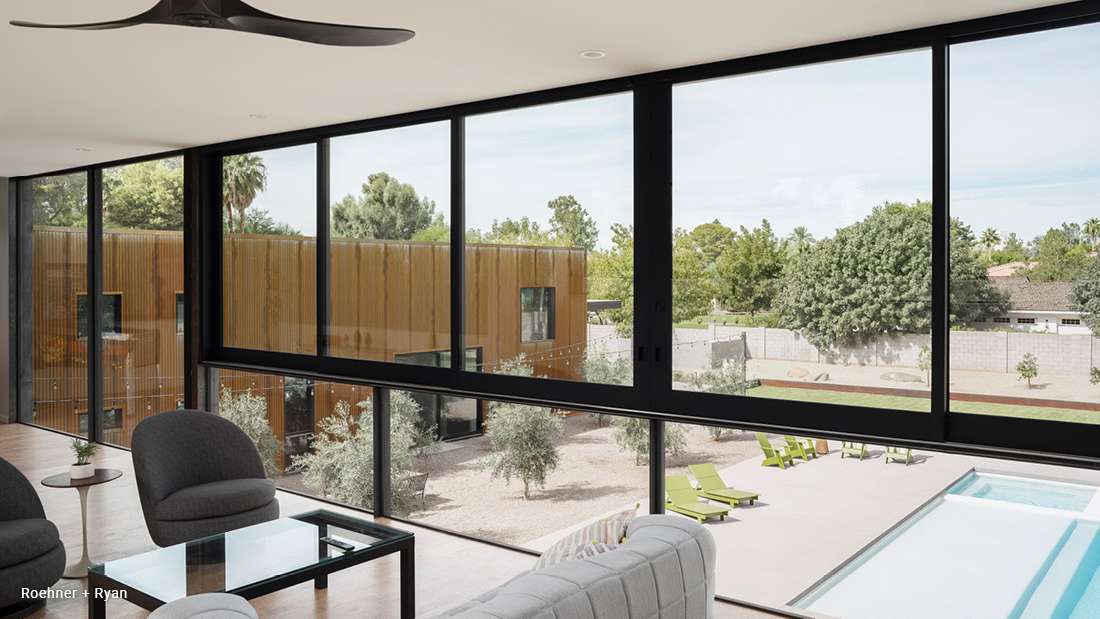 Modern living room with floor-to-ceiling black-framed windows overlooking a backyard with swimming pool, green lounge chairs, and a contemporary wood-clad building.