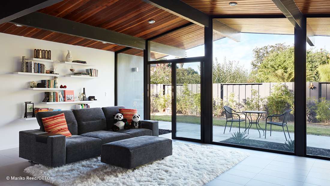 Cozy modern living room with floor-to-ceiling glass doors opening to a patio, wooden ceiling beams, gray sectional sofa, and minimalist wall shelving.