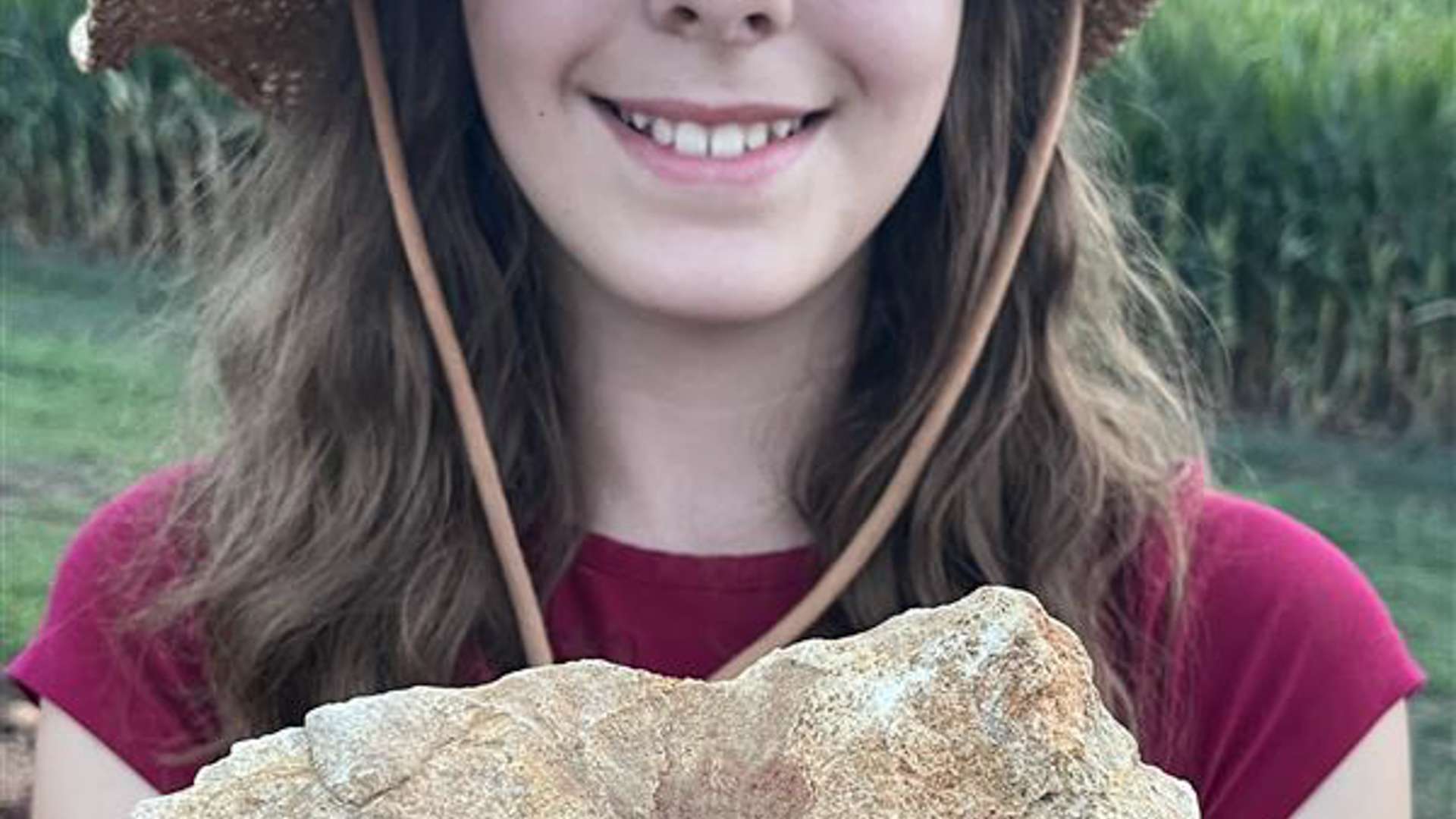 Person wearing a straw hat and red shirt holding a large, rough-textured tan rock with both hands, standing outdoors in front of a green cornfield.