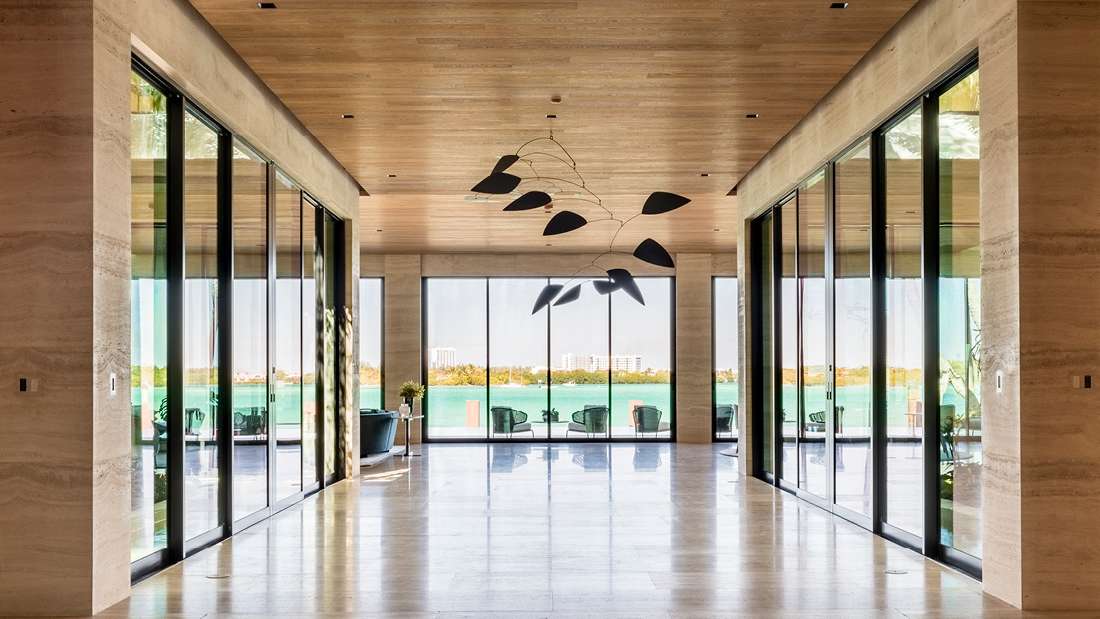 Luxury modern hallway with floor-to-ceiling glass doors on both sides, natural stone walls, light wood ceiling, and panoramic waterfront view framed by large sliding glass panels, featuring sculptural black mobile chandelier.