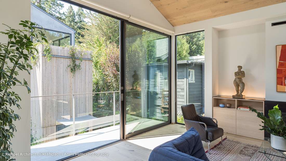 Modern sitting area with floor-to-ceiling glass doors, natural wood ceiling, contemporary armchairs, and views of a lush garden.
