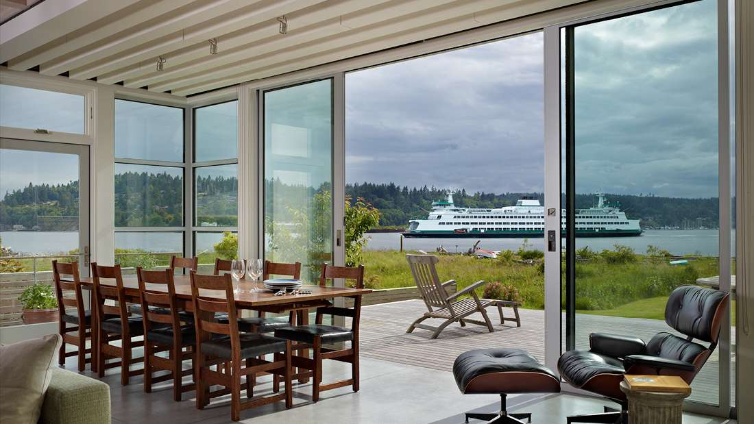 Modern dining area with wooden table and chairs, floor-to-ceiling glass walls offering waterfront views, and outdoor deck with lounge chairs overlooking a passing ferry.