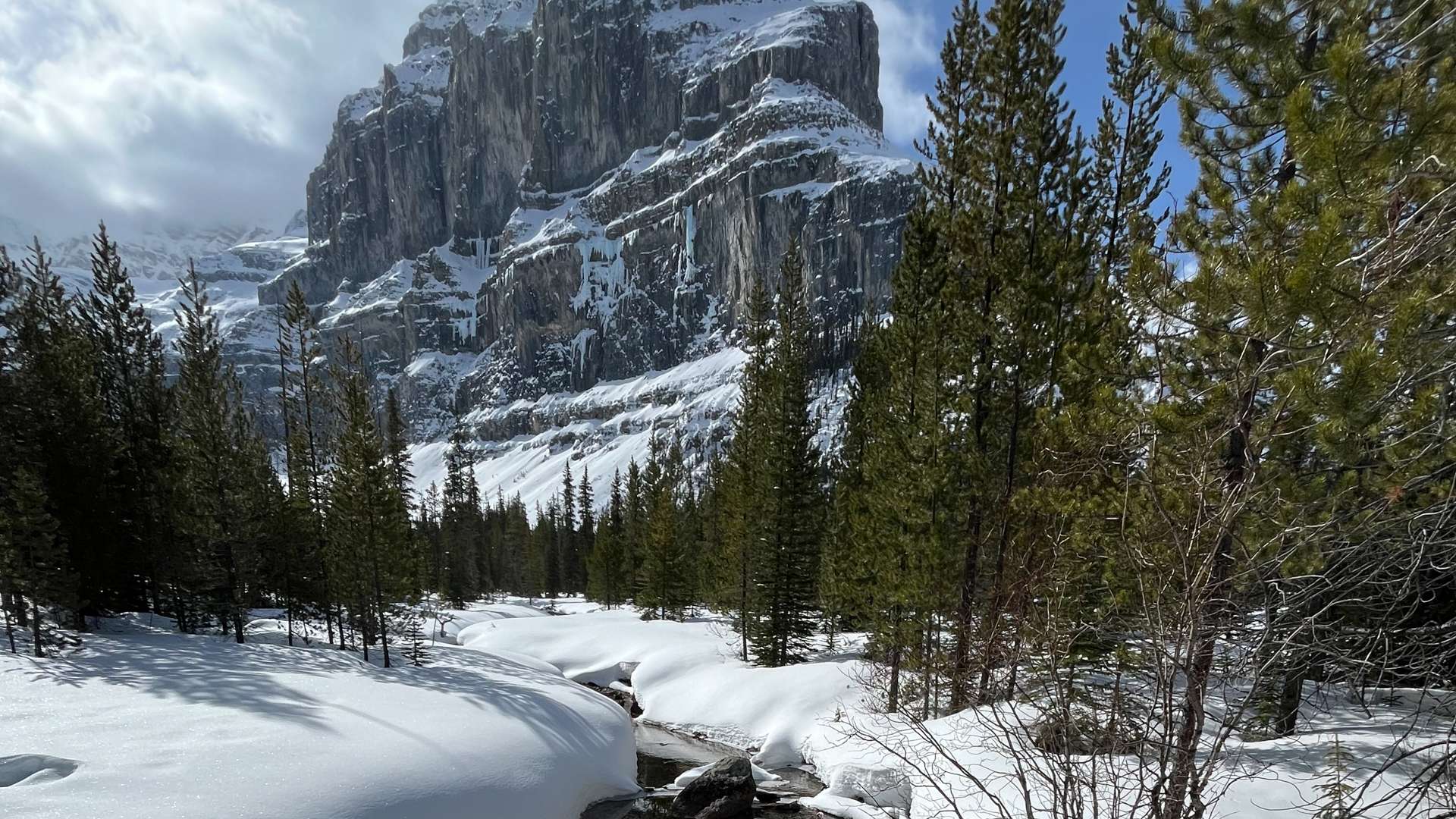 Towering rocky mountain with snow-covered slopes, surrounded by evergreen trees and a narrow stream under a partly cloudy sky.
