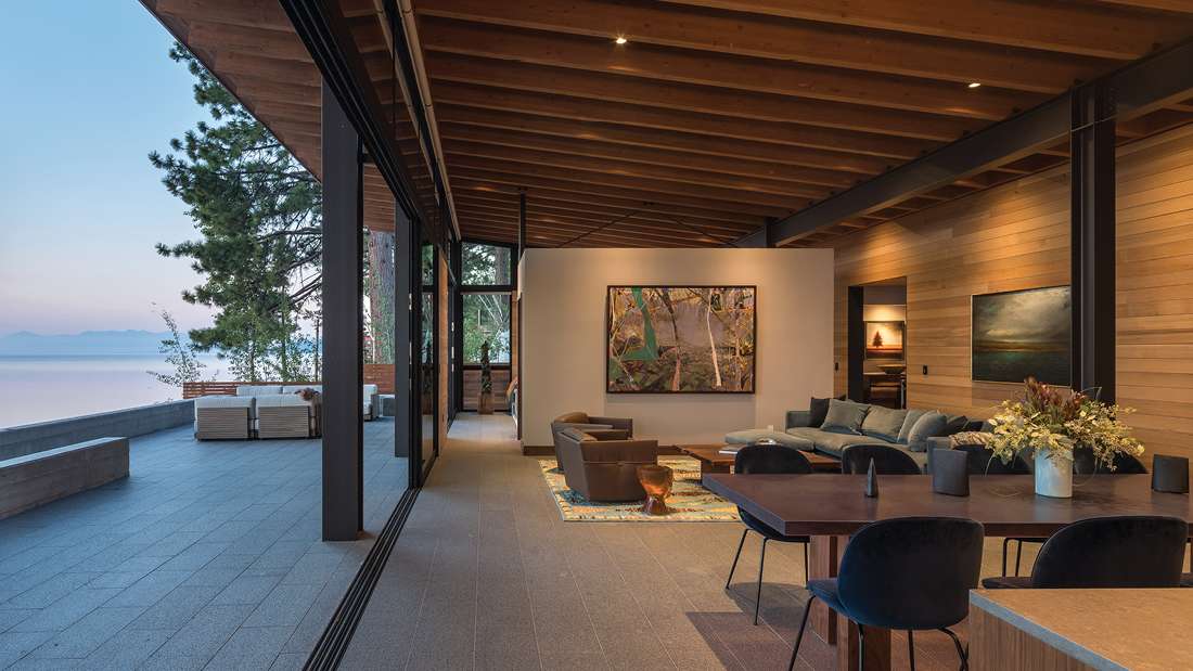 Luxury lakefront living space with exposed wood ceiling beams, floor-to-ceiling glass walls, open-concept layout featuring modern dining table, plush sectional sofa, and outdoor patio overlooking serene water views.