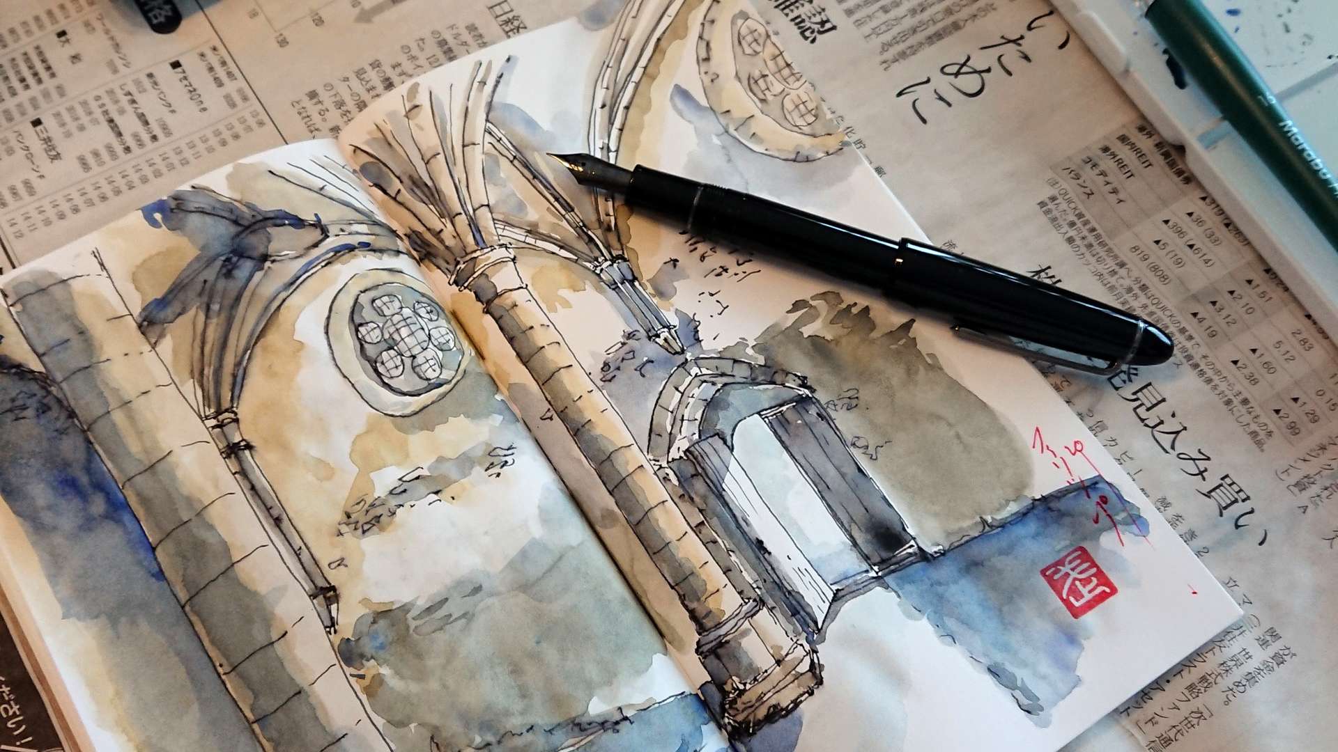   Watercolor sketch of stone arches in an open notebook with a fountain pen, paintbrush, and palette on a newspaper.