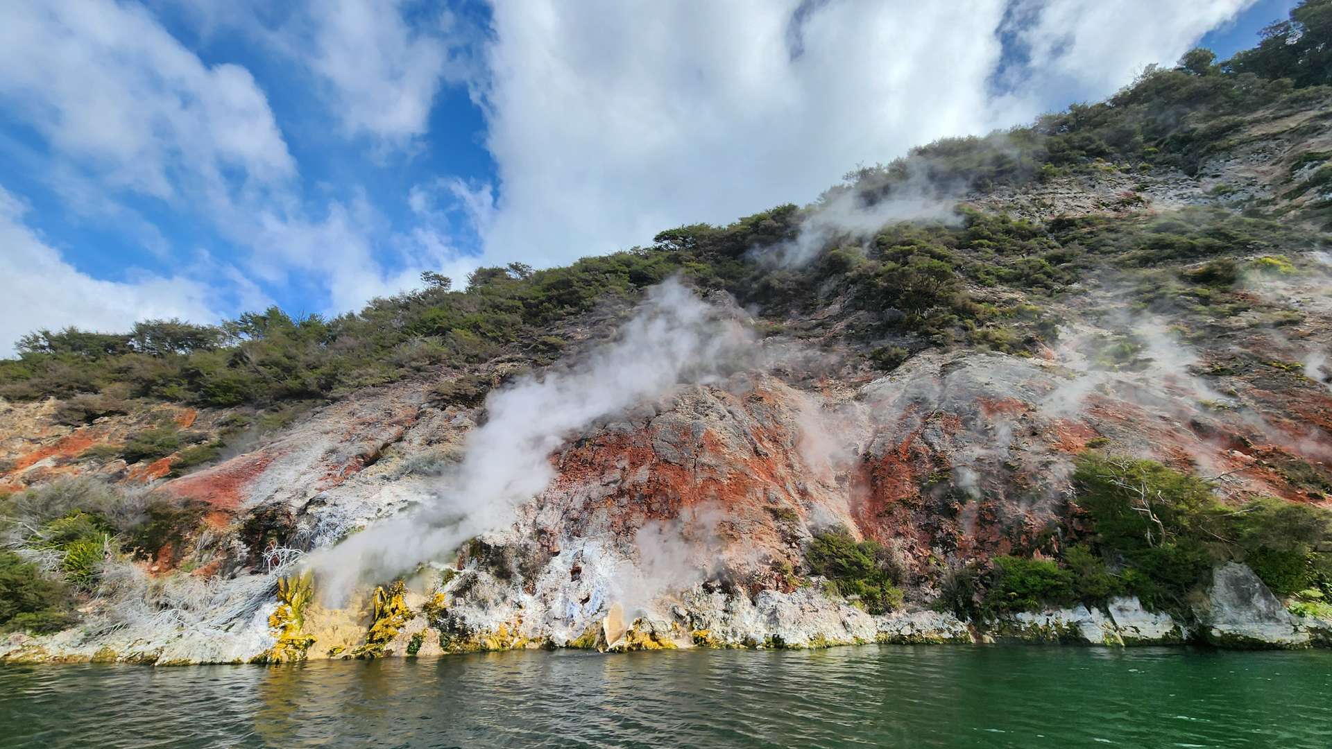 Steaming geothermal hillside with red and yellow mineral deposits above a green lake under a partly cloudy sky.