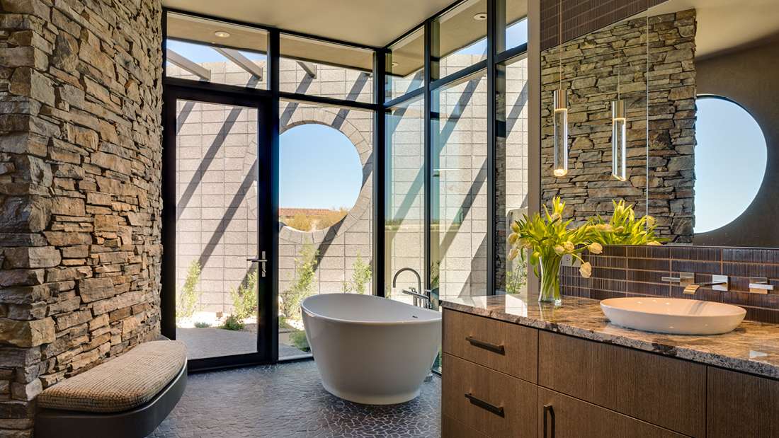 Luxury bathroom with freestanding white soaking tub, stone accent walls, large glass doors opening to private courtyard, and modern vanity with granite countertop and vessel sink.