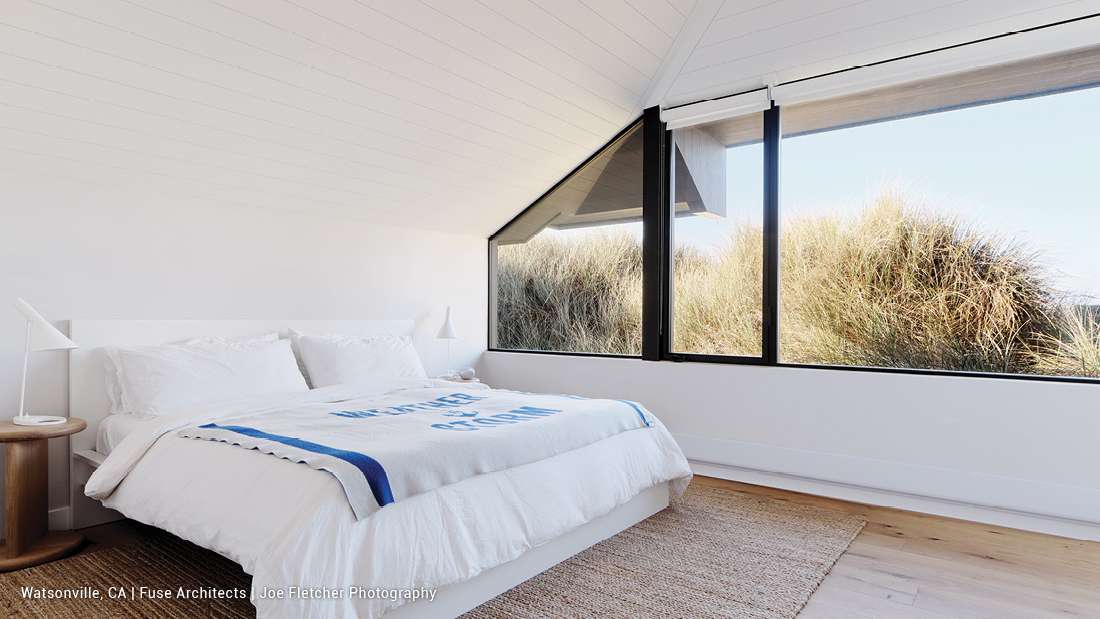 Minimalist coastal bedroom with white bed frame, crisp white bedding, light wood side table, modern white lamp, and large black-framed windows overlooking sand dunes and natural grasses.