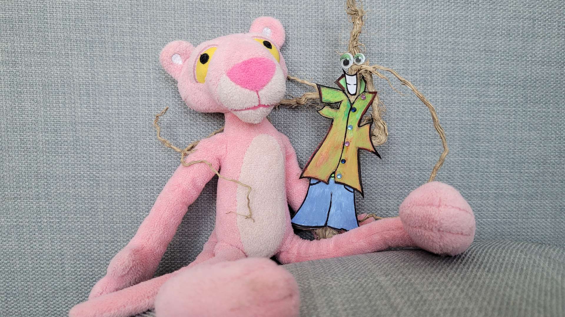 Pink Panther plush toy sitting on a gray fabric surface, holding a colorful paper cutout figure with googly eyes and twine arms, dressed in a green coat and blue pants.