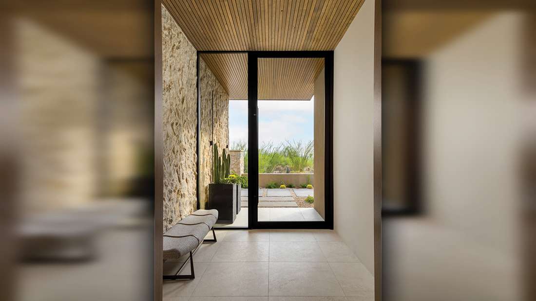 Minimalist entryway with black-framed glass pivot door, textured stone accent wall, light beige tile flooring, and wood slat ceiling leading to an outdoor patio with desert landscaping.