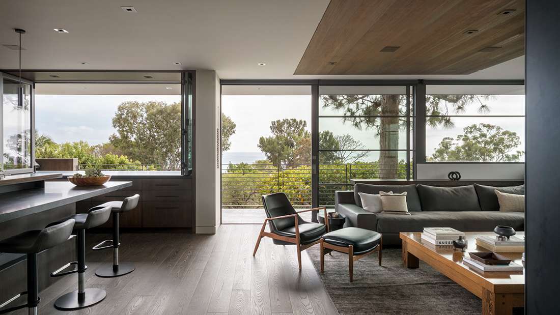 Modern open-concept living room with floor-to-ceiling glass doors opening to a balcony, gray sectional sofa, wooden coffee table with books, black leather lounge chair, and kitchen bar with black stools overlooking ocean and tree views.