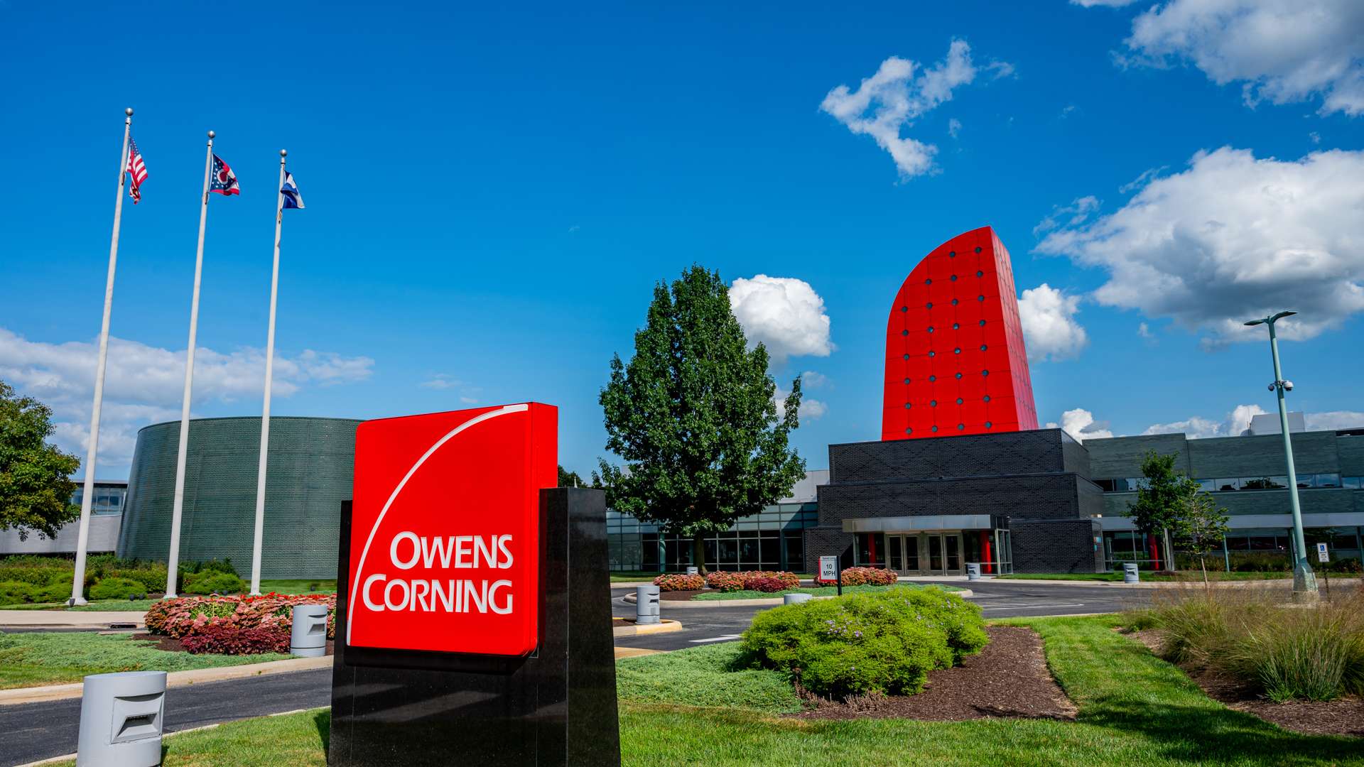 Owens Corning headquarters with red architectural feature, company sign, and flagpoles under a bright blue sky.