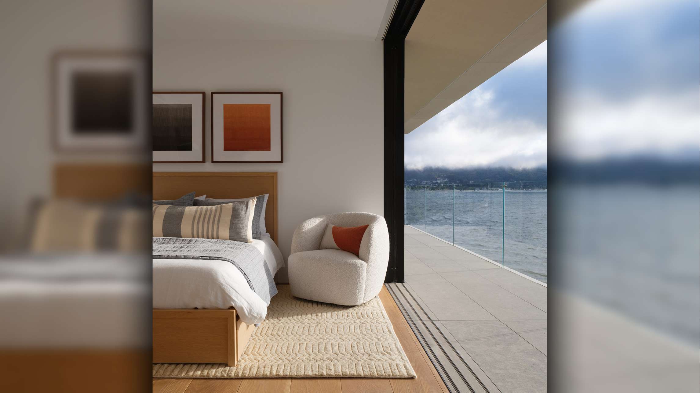 Modern bedroom with minimalist decor, light wood bed frame, white bedding with gray and beige accent pillows, textured cream area rug, and large sliding glass doors opening to a balcony with panoramic ocean views.