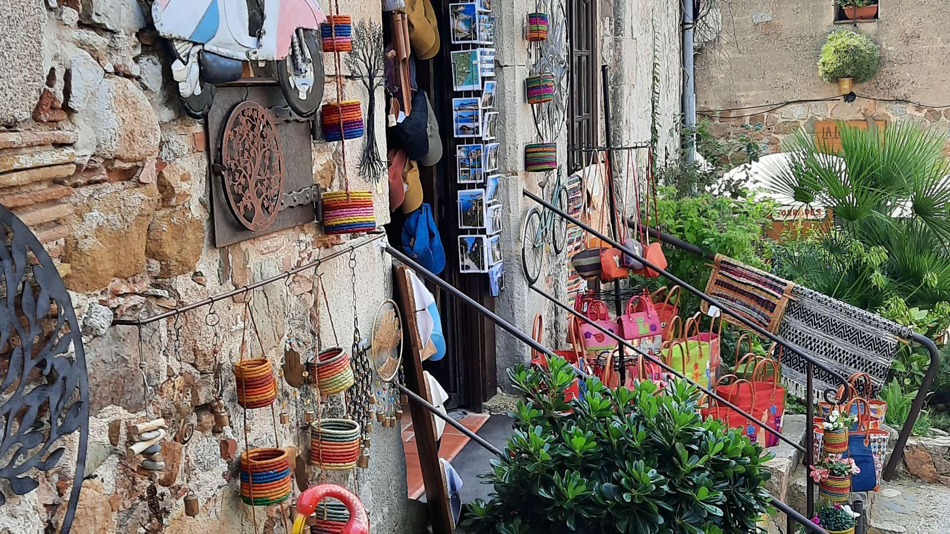 Rustic shop with colorful handmade crafts, metal art, and plants on a stone wall and steps.